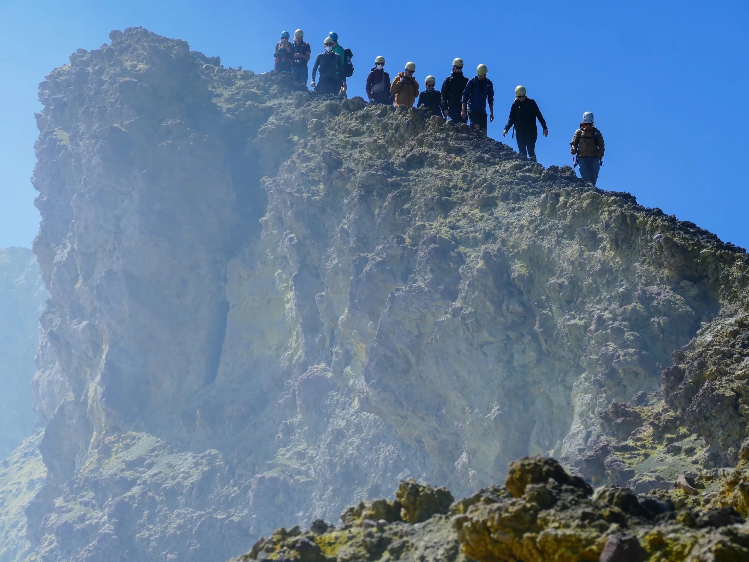 Escursionisti con casco in fila sul bordo di un cratere sommitale dell'Etna, tra rocce gialle di zolfo